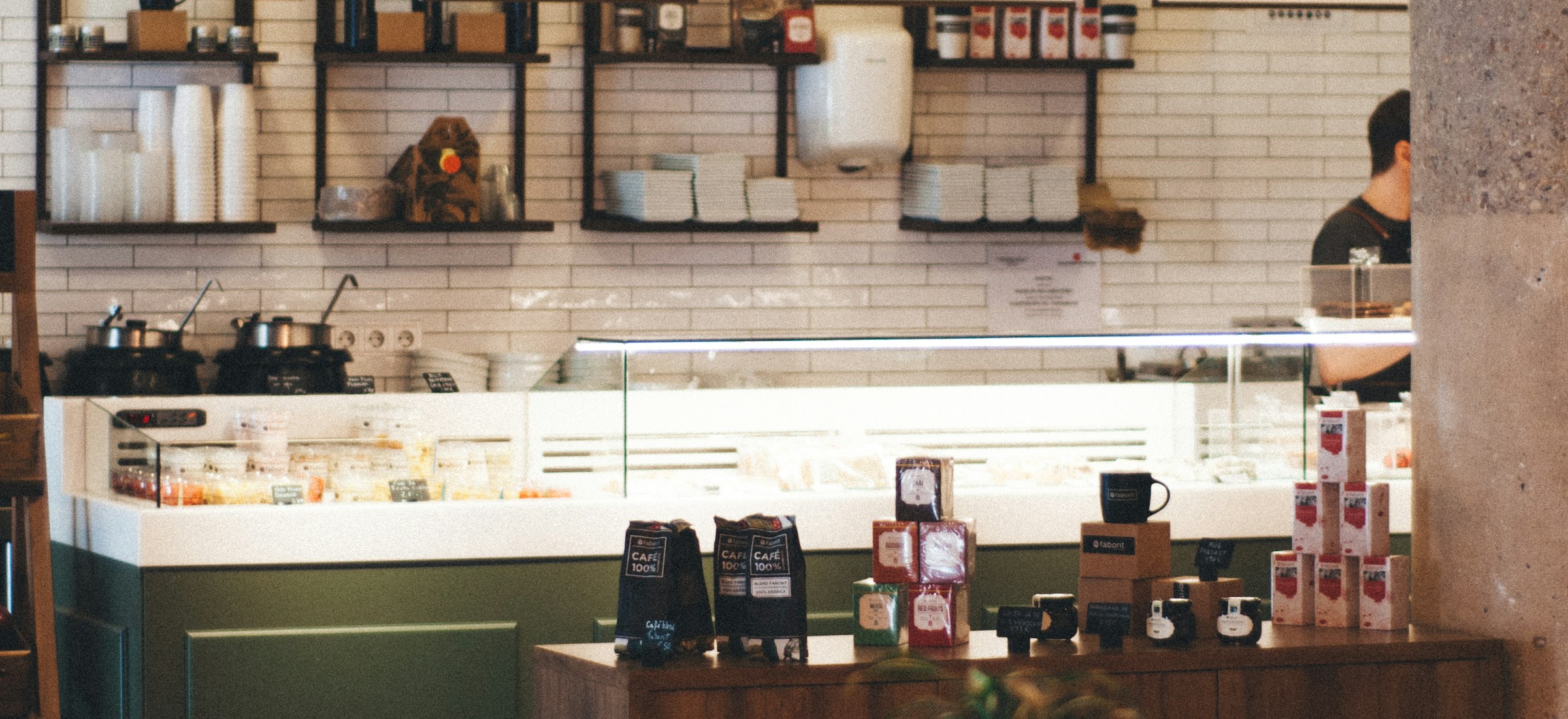 A warm neighborhood cafe interior: tiled counter, hand-lettered menu boards, and a row of pendant lights over a long communal table